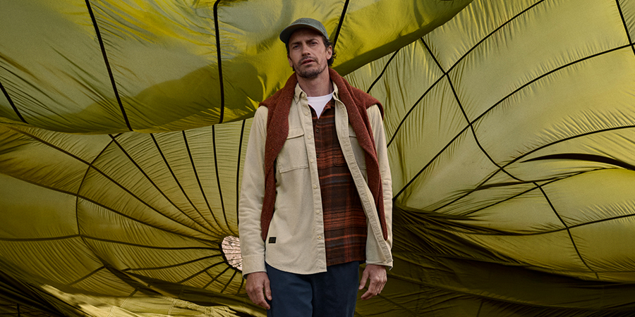 Mann in beiger Jacke und kariertem Hemd vor einem gelb-grünen, gewellten Hintergrund.
