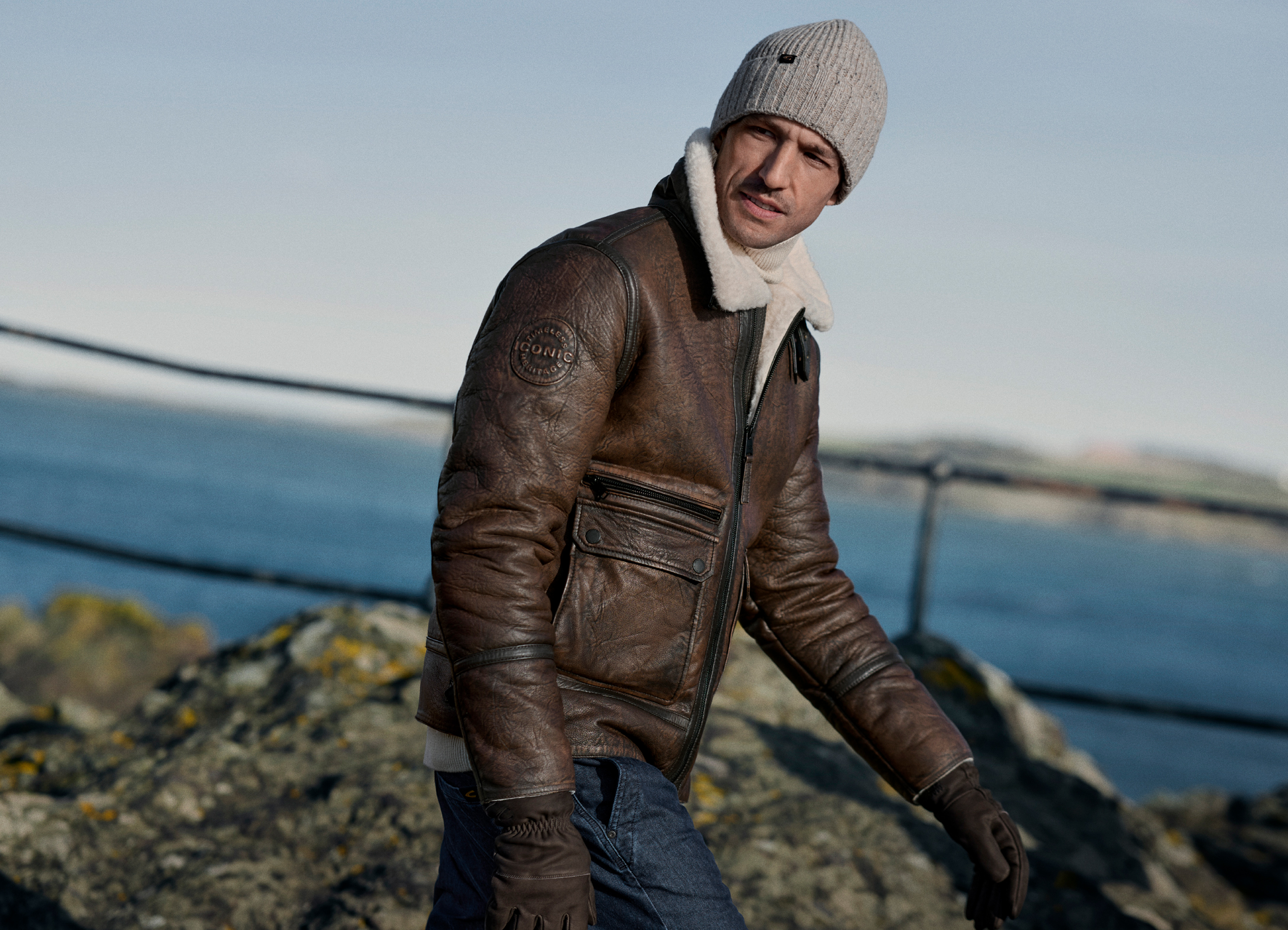 Man in a brown leather jacket and grey beanie stands by the coast with the sea in the background.