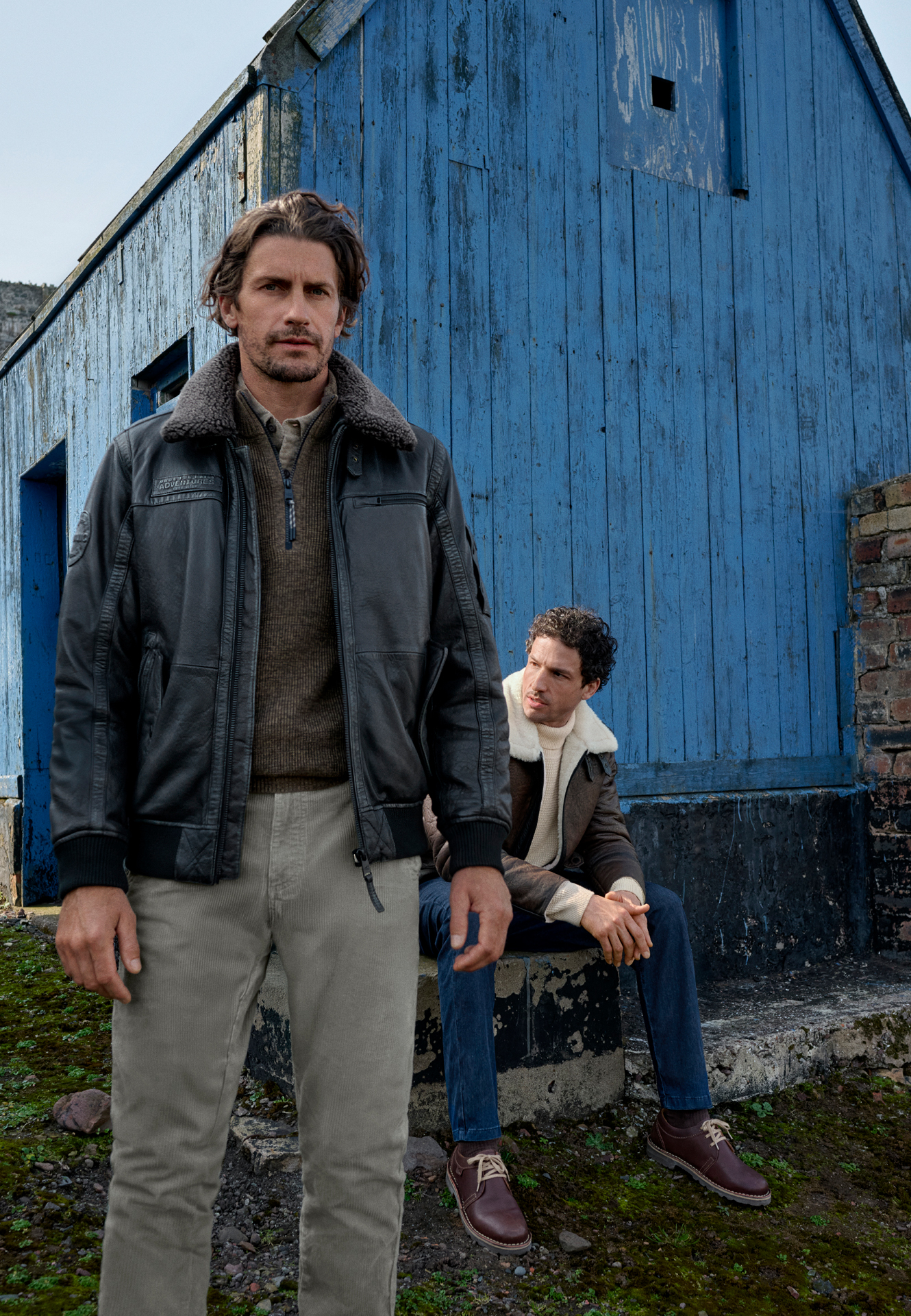 Two men in stylish clothing in front of a blue wooden cabin; one is wearing a leather jacket.