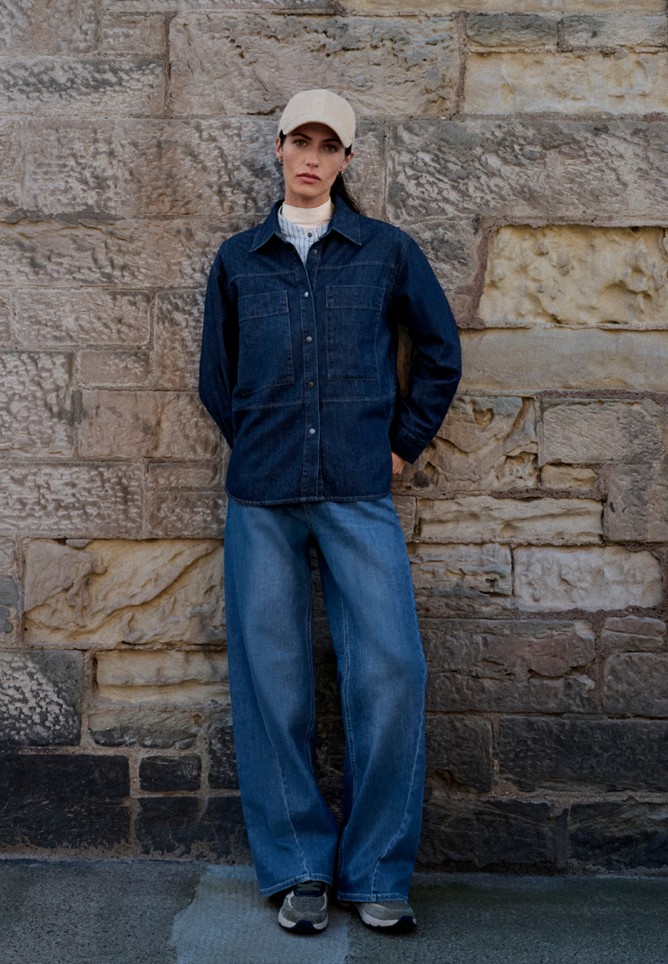 Eine Frau trägt eine dunkelblaue Jeansbluse und weite Jeans vor einer steinernen Wand.