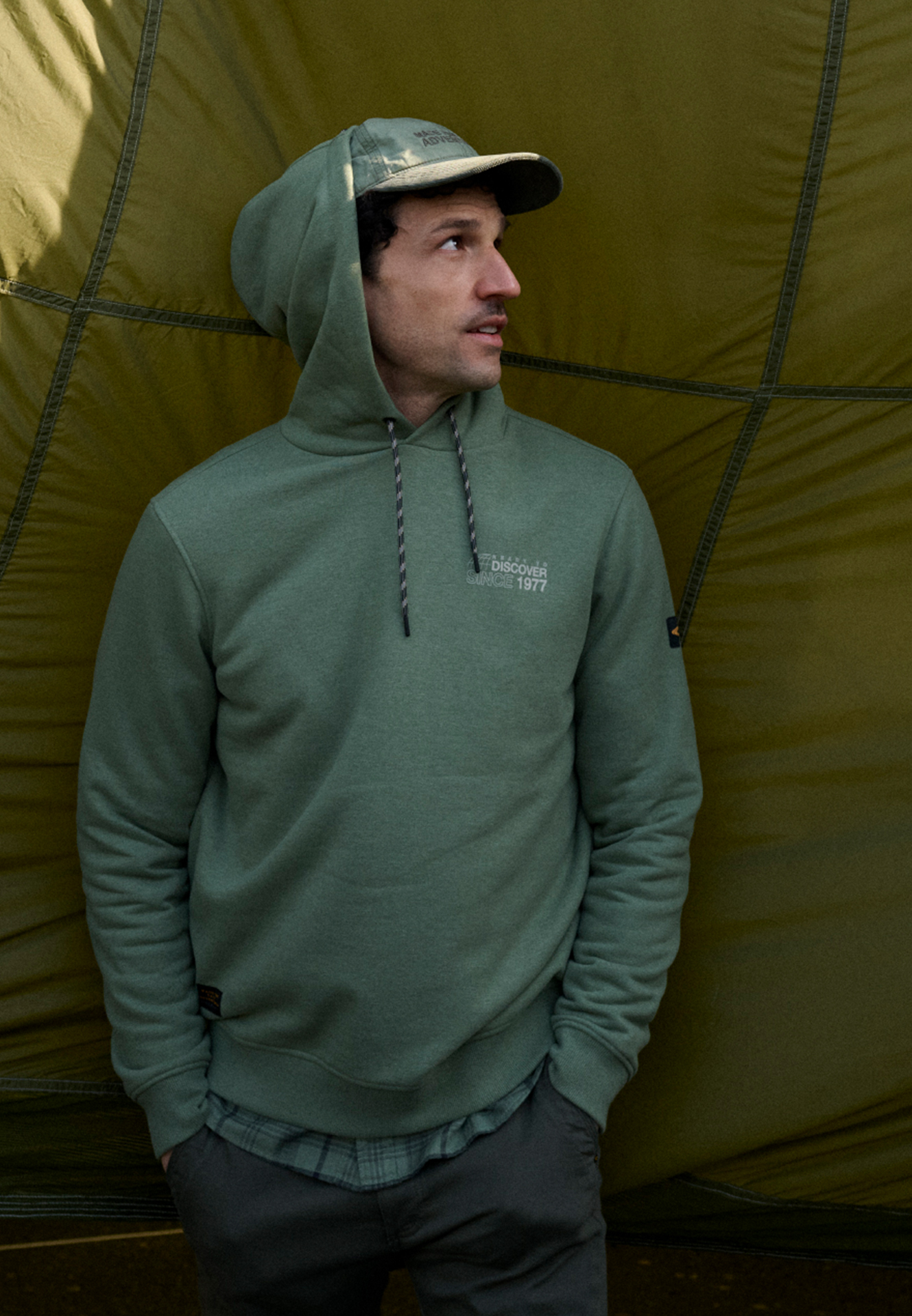 Man in zwarte hoodie en camouflage pet voor een groene tentwand.