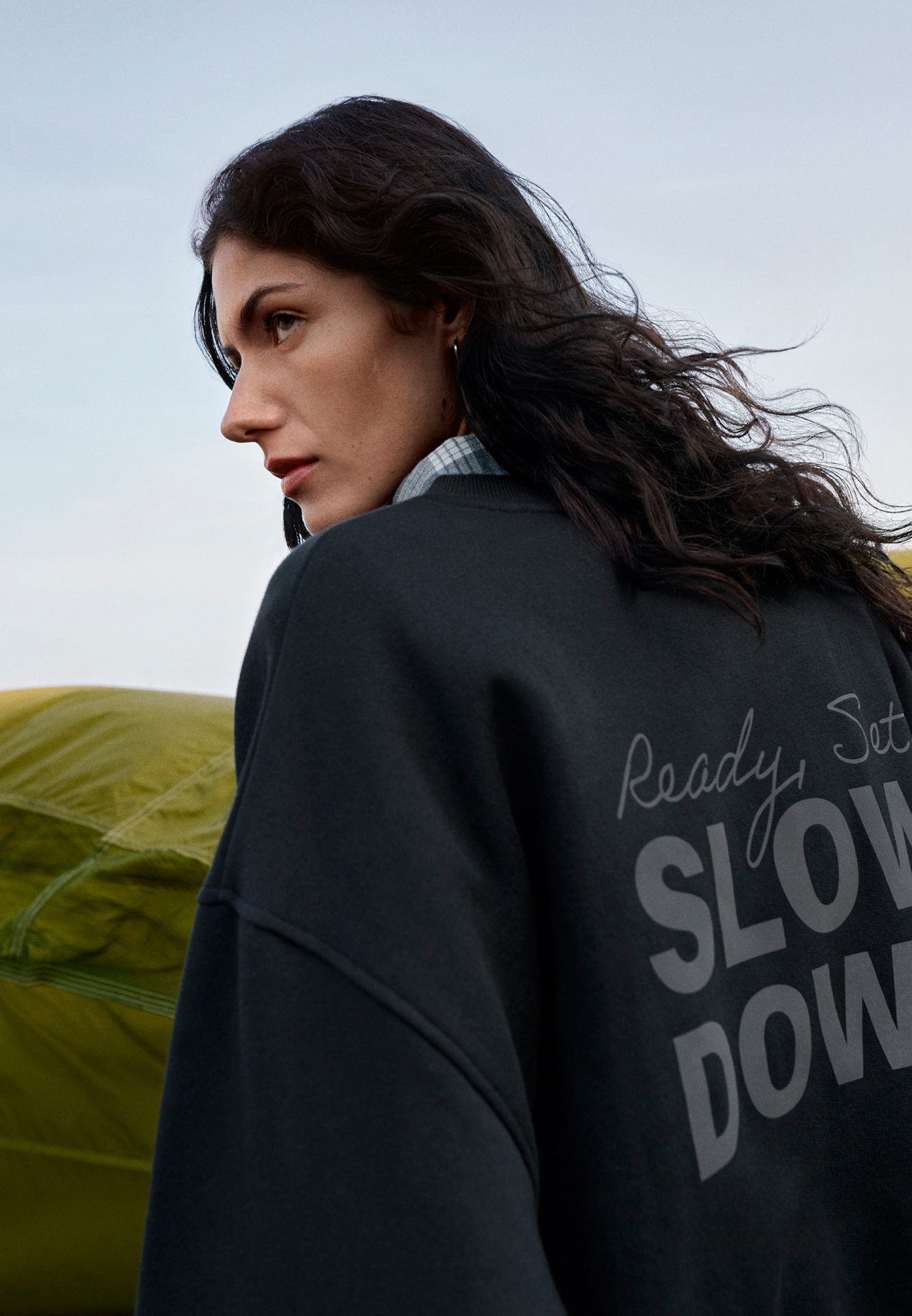 Een vrouw met lang haar draagt een zwart sweatshirt met de tekst 'SLOW DOWN' buiten.