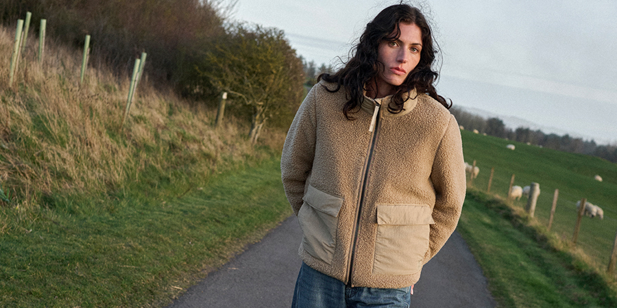 Eine Frau mit langen, dunklen Haaren trägt eine beige Teddyjacke und steht auf einem Feldweg.