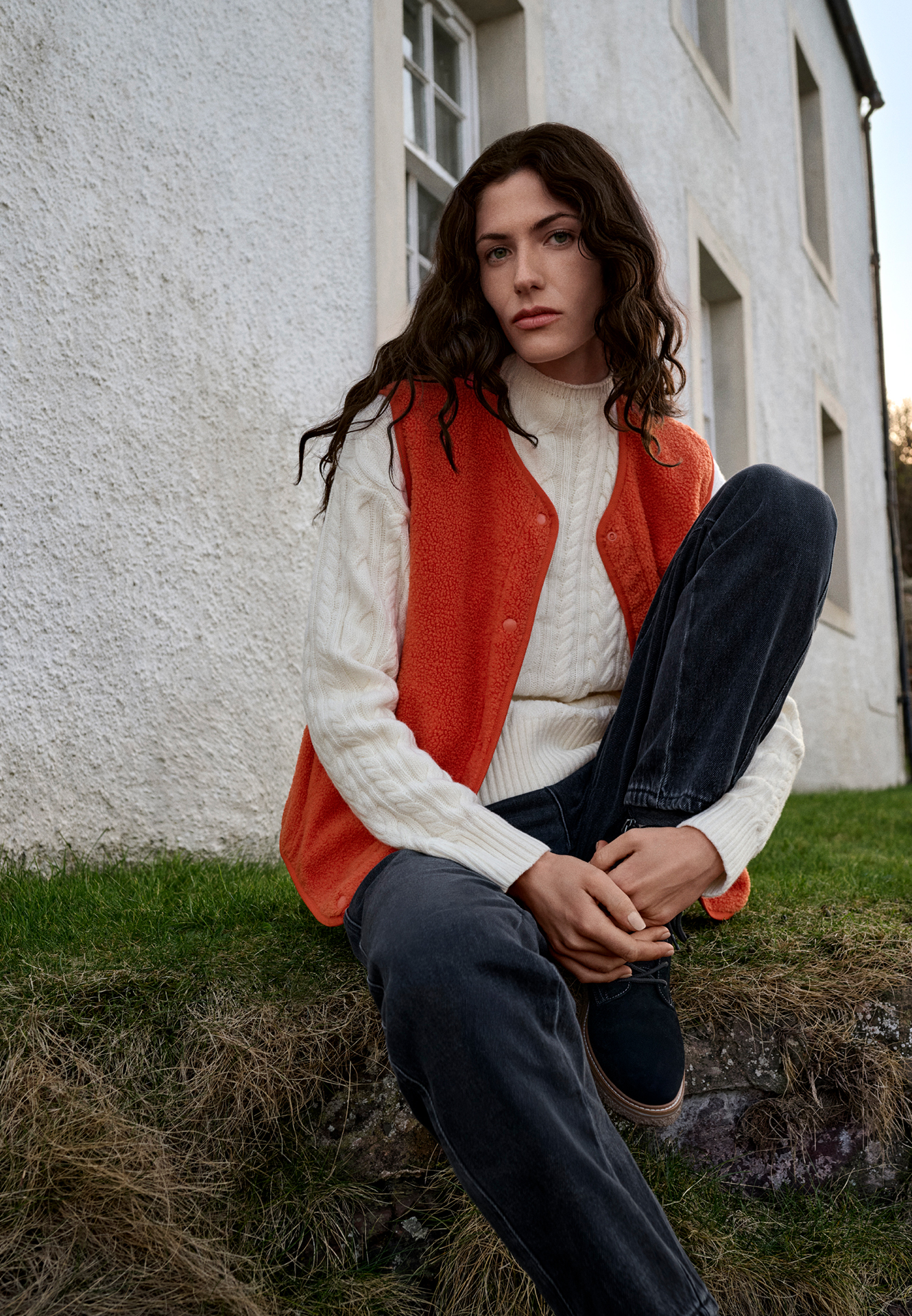 Jonge vrouw in een oranje teddyvest en witte trui zit op het gras voor een gebouw.