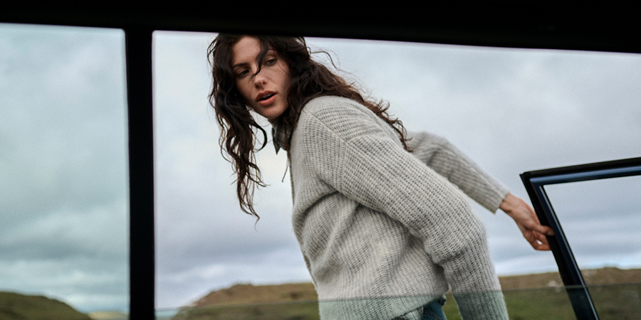 Eine Frau mit lockigem Haar trägt einen grauen Strickpullover und steigt aus einem Fahrzeug.